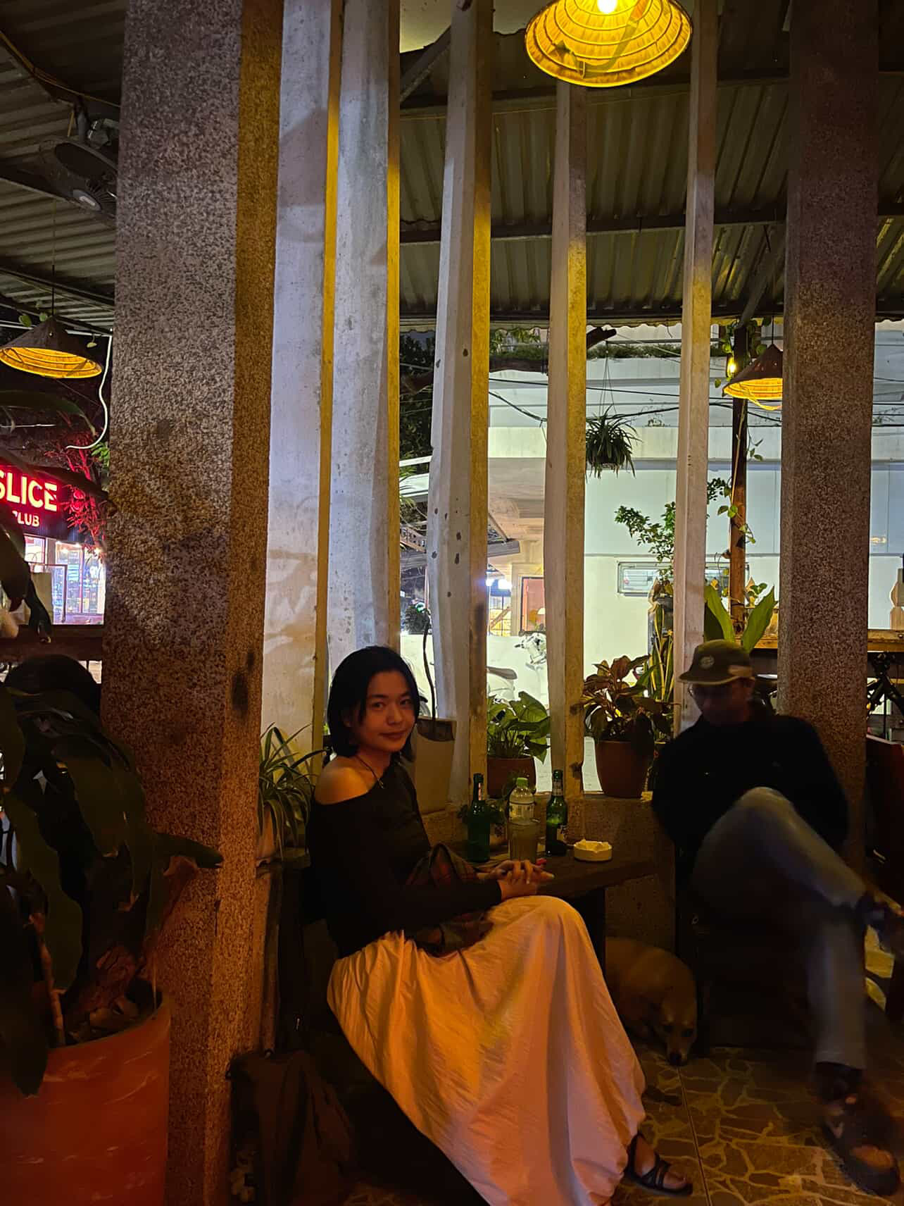 Woman seated between concrete columns at night, neon visible through the window